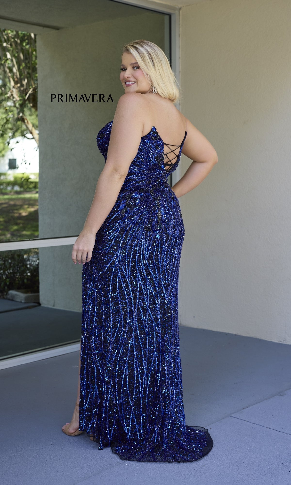 A woman stands outdoors near a window, wearing the Plus-Size Strapless Prom Gown: Primavera 14083—a stunning floor-length blue sequin dress with a lace-up back, perfect for any special occasion.